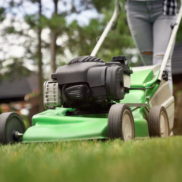 Close up of female legs of gardener using green lawn mower on backyard. Selective focus of woman working in summer, cutting grass in backyard. Concept of gardening, work, nature.